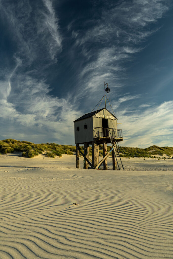 Het iconische drenkelingenhuisje op het strand nabij paal 19 op Terschelling, foto genomen vanaf het strand, tevens duin en sluierbewolking te zine tegen een blauwe lucht. Terschelling © Marjolein van Roosmalen