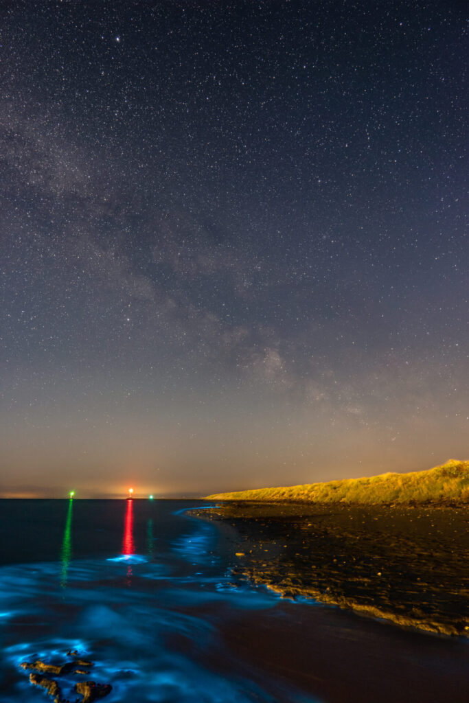 Een heldere nachtelijke lucht met sterren van de melkweg boven het donkere water in de baai van West-Terschelling. In de branding oplichtend zeevonk en in de verte navigatiebelichting van de betonning. © Marjolein van Roosmalen Terschelling